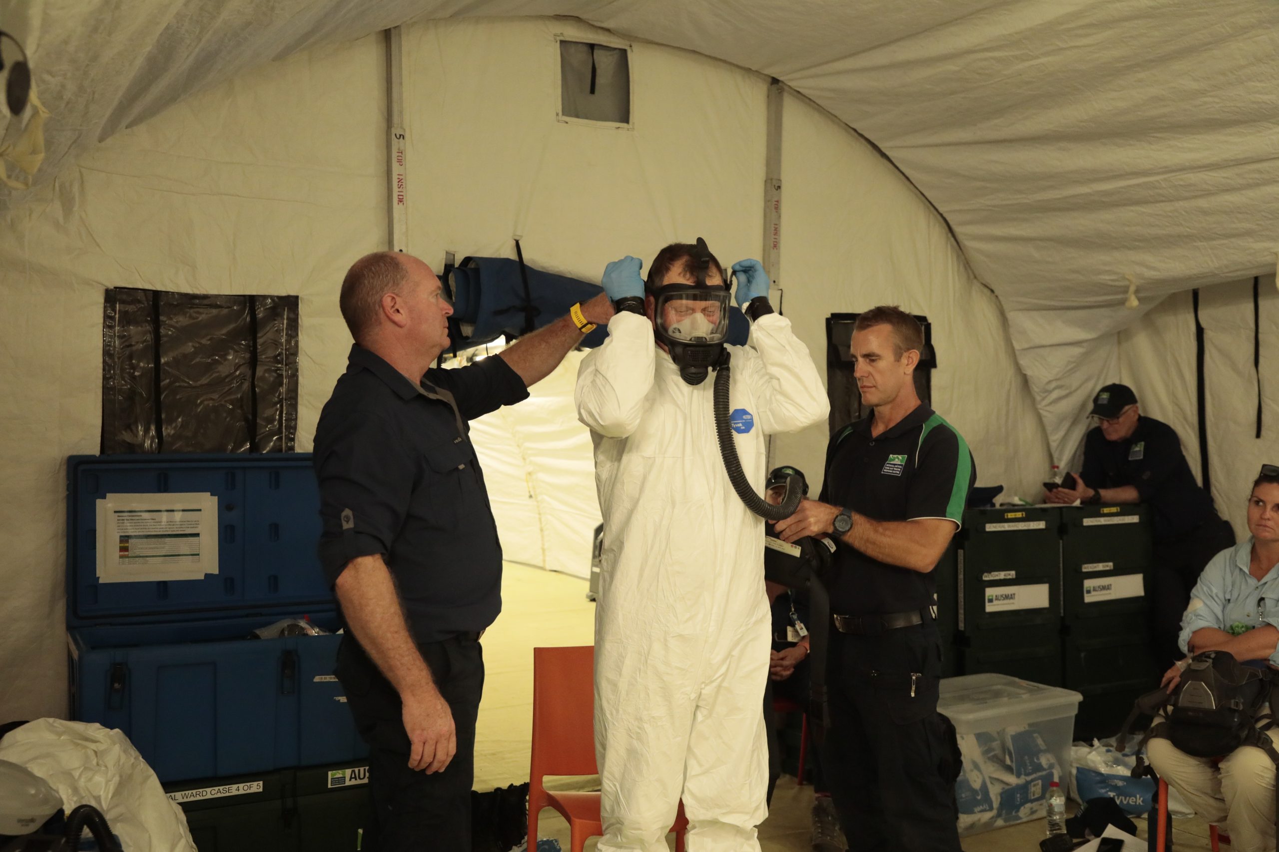 A person in a white suit with a mask and two persons assisting.